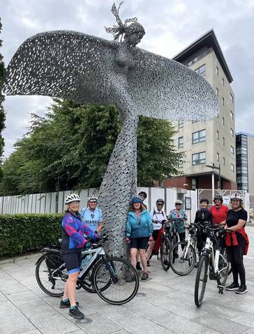 Female cyclists posing in front of large iron angel sculpture.