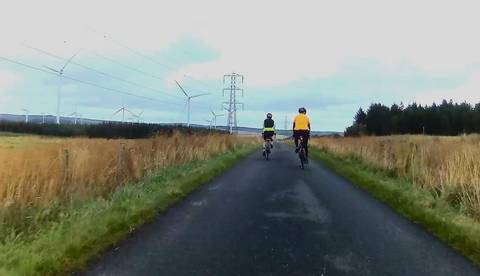 Two cyclists on tarmac road alongside multiple wind turbines.
